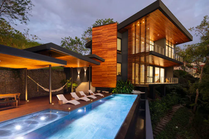 Modern luxury home with infinity pool, lounge chairs on wooden deck, surrounded by lush tropical greenery at dusk