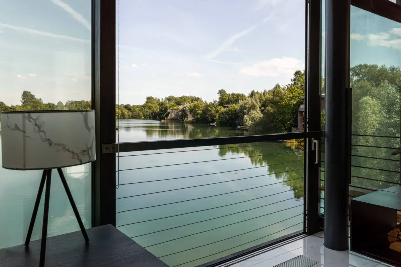 Modern glasshouse interior with marble lamp on table, overlooking serene lake and lush green trees.