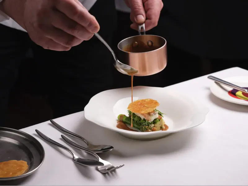 Chef pouring sauce from copper pan onto plated dish with greens and orange crisp on white tablecloth