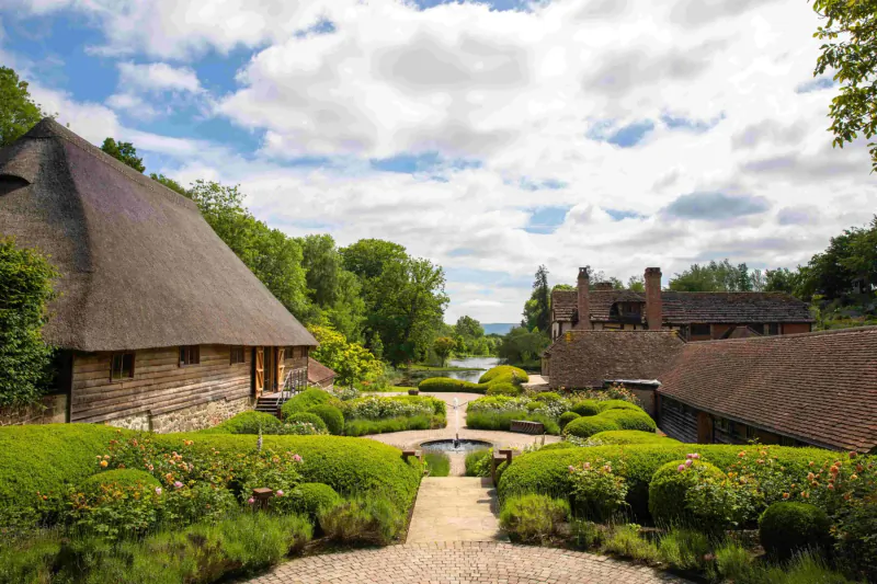 Thatched-roof cottages and lush gardens with fountain path, under partly cloudy sky