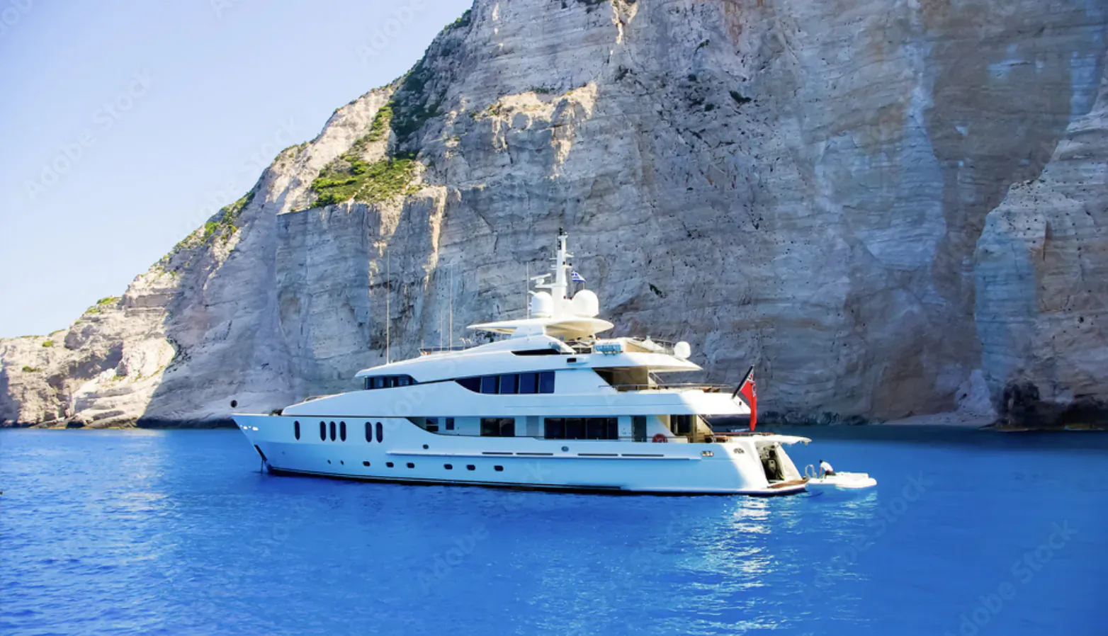 White luxury yacht with red flag anchored in blue waters near steep cliffs of Zakynthos, Greece.