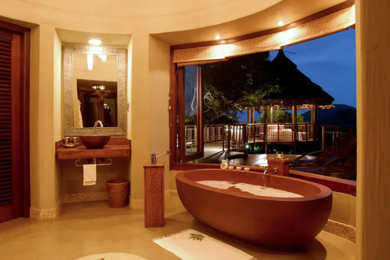 Luxurious safari lodge bathroom with freestanding terracotta bathtub, wooden vanity, and sunset view of thatched deck.