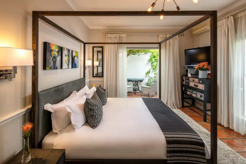 Spacious bedroom in Spanish Farm Guest Lodge with four-poster bed, modern art, TV, and balcony view of garden patio.
