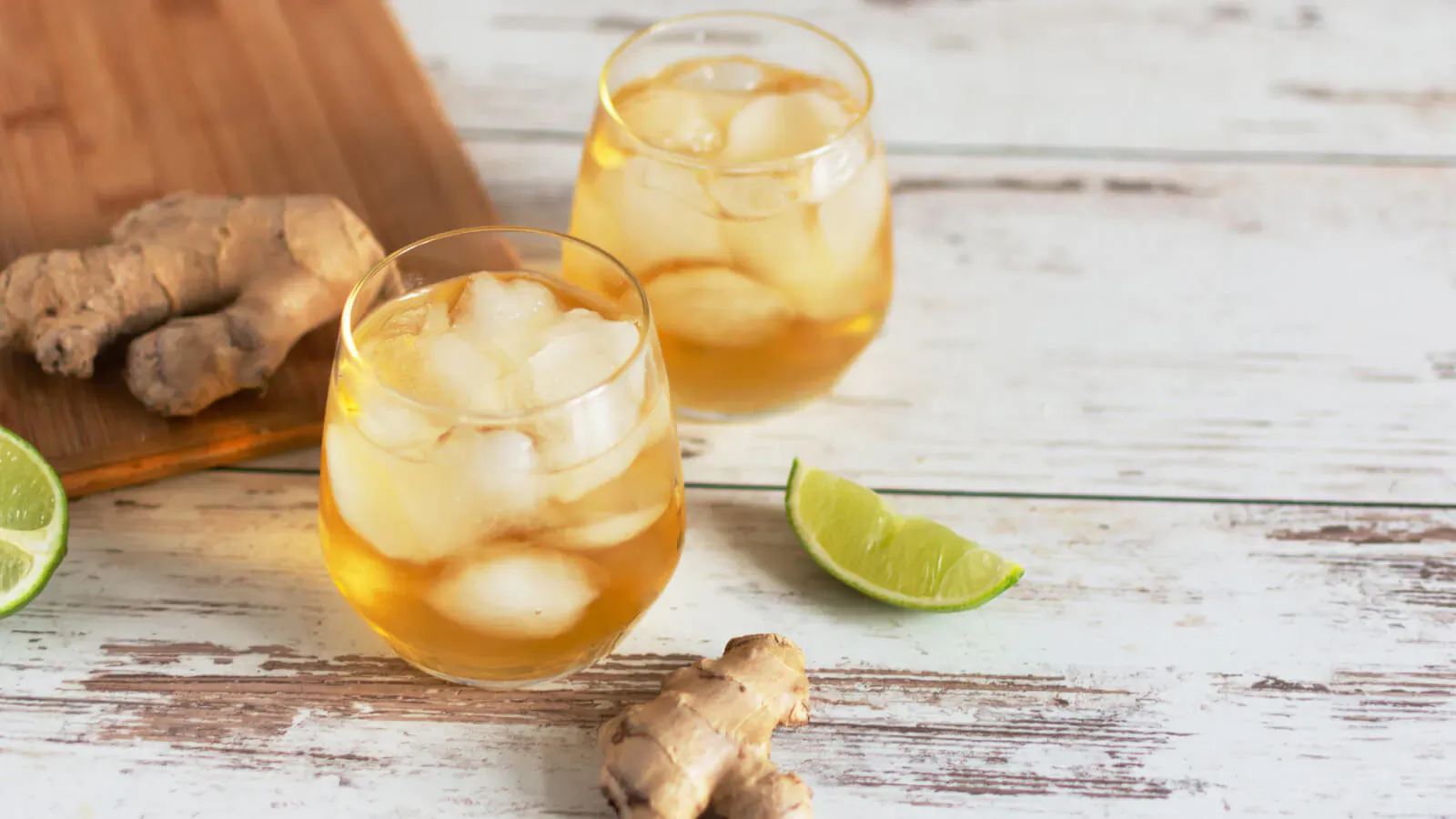 Two glasses of iced amber Gunner Saint mocktail with ginger root and lime slices on wooden surface.