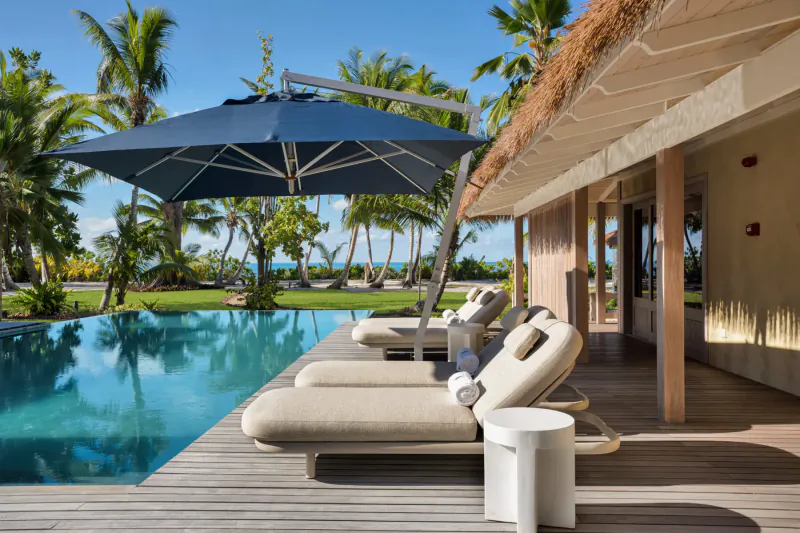 Luxury poolside deck at Four Seasons Desroches villa with beige loungers, blue umbrella, thatched roof, and tropical beach backdrop.