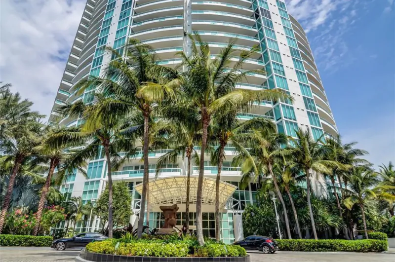 Murano Grande luxury high-rise entrance with glass canopy, palms, landscaping, and cars under blue sky.