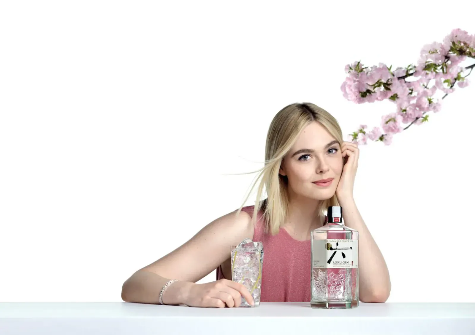 Elle Fanning smiles, hand on cheek, holding Roku Gin bottle and glass with pink cherry blossoms on white background