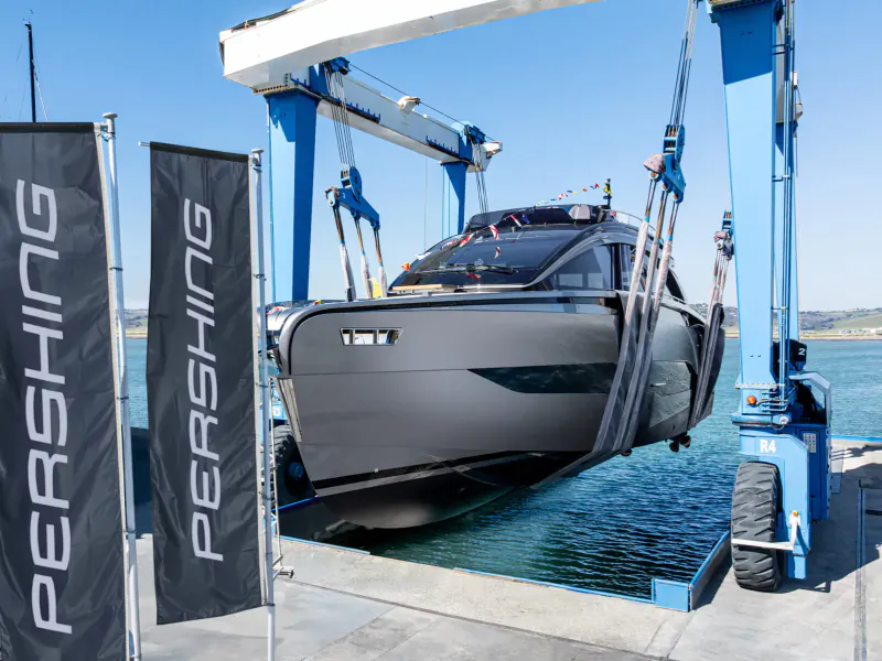 First Pershing GTX70 yacht lifted by blue crane from water near black Pershing flags, sunny day.