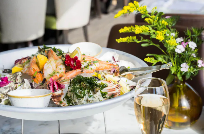 Large white platter of shrimp, lemon slices, edible flowers, and rice, garnished with herbs, beside champagne glass and flowers on table.