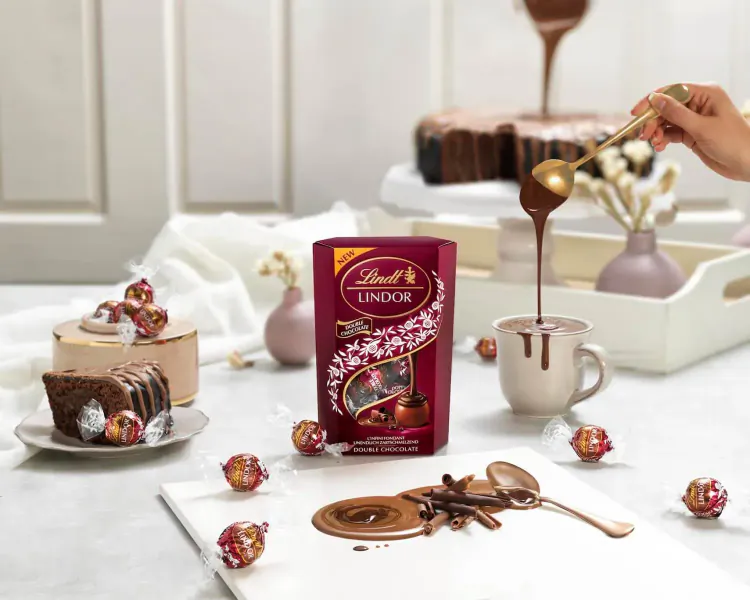 Lindt Lindor chocolate box with truffles, cake slices, and molten chocolate poured from spoon on table.