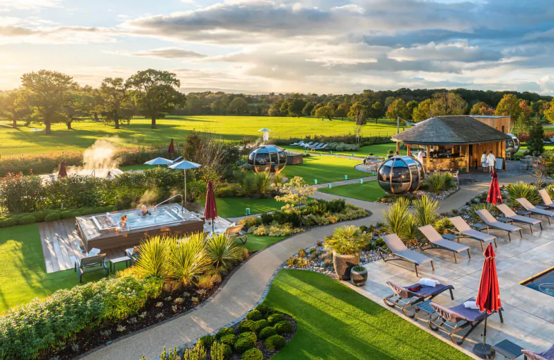 Aerial view of Carden Park spa garden at sunset with hot tub, pool, loungers, umbrellas, fountain, and lush lawns.