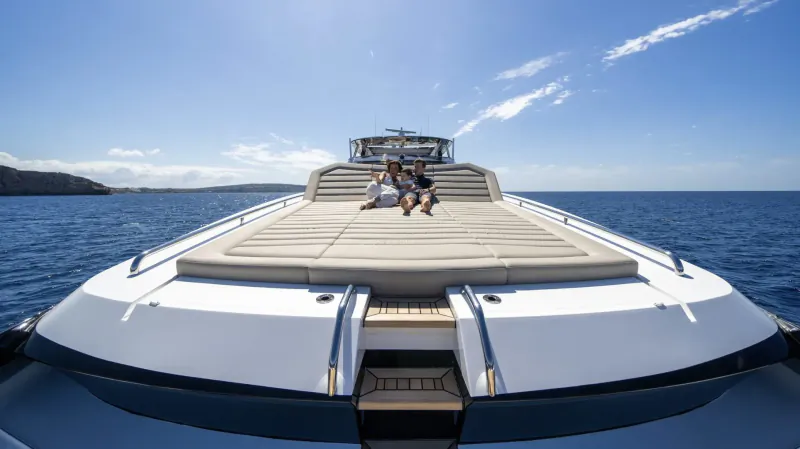 Front view of white luxury yacht with two people seated on sun deck, blue sea and cliffs ahead under clear sky.