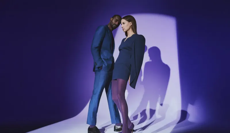 Black man in blue suit and woman in navy dress with purple stockings stand intimately under purple spotlight with shadows, Falke gallery.