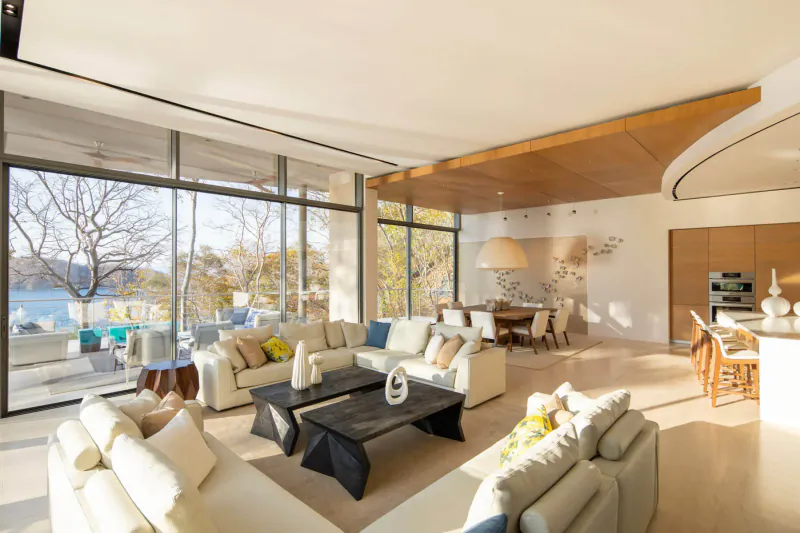 Spacious modern luxury living room with white sofas, black coffee table, open kitchen, and panoramic lake views through large windows.
