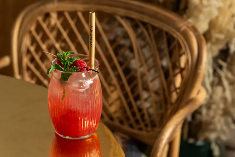 Pink cocktail with raspberry, mint, and gold straw in tall glass on gold table, rattan chair backdrop