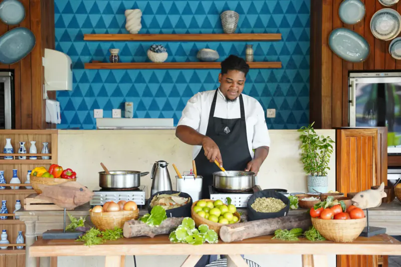 Smiling chef in white uniform and black apron stirring pot amid fresh fruits, veggies on wooden table in modern kitchen with blue geometric wall.