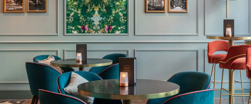 Elegant bar interior at Browns Cardiff with teal velvet chairs, gold tables, candles, and green floral wall art.