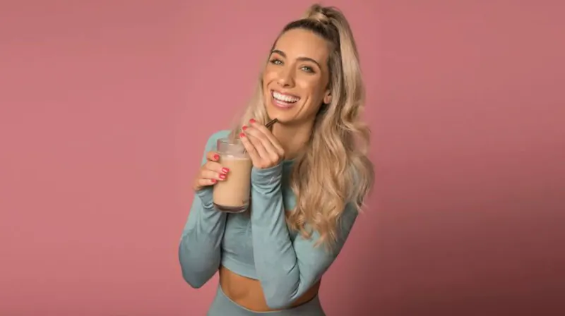 Blonde woman in ponytail and teal crop top smiling, holding pink smoothie on pink background