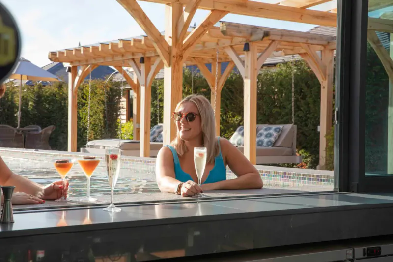 Blonde woman in turquoise bikini and sunglasses sits at poolside bar at The Manor House Hotel & Spa Cheshire, holding champagne flute amid cocktails.
