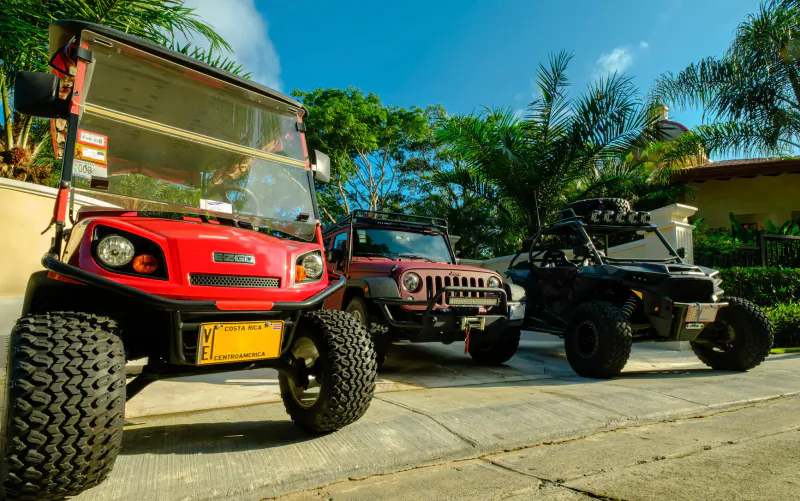 Red golf cart, Jeep Wrangler, and black dune buggy parked on driveway amid tropical palms at luxury villa
