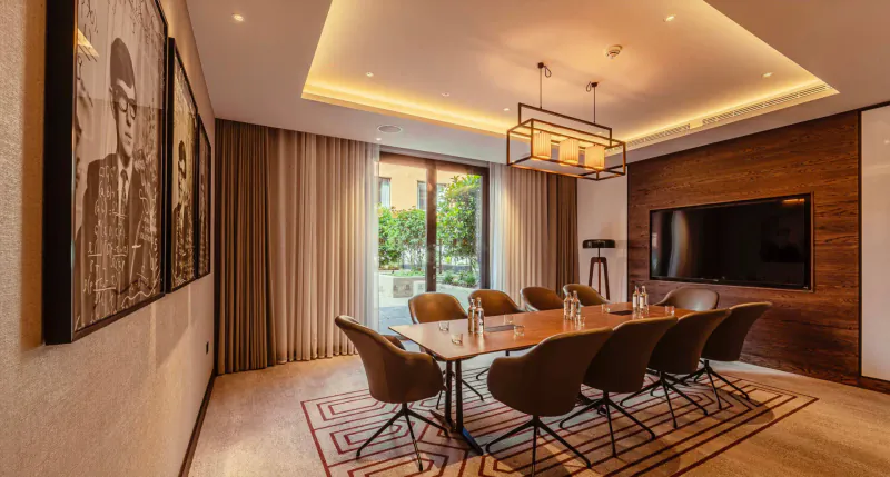 Elegant meeting room in luxury apartment with long wooden table, swivel chairs, pendant lights, TV, and black-and-white portraits overlooking garden.