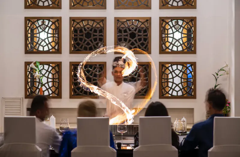 Chef twirling flames in heart shape at elegant dining table with guests, ornate wall screens, Maldives resort