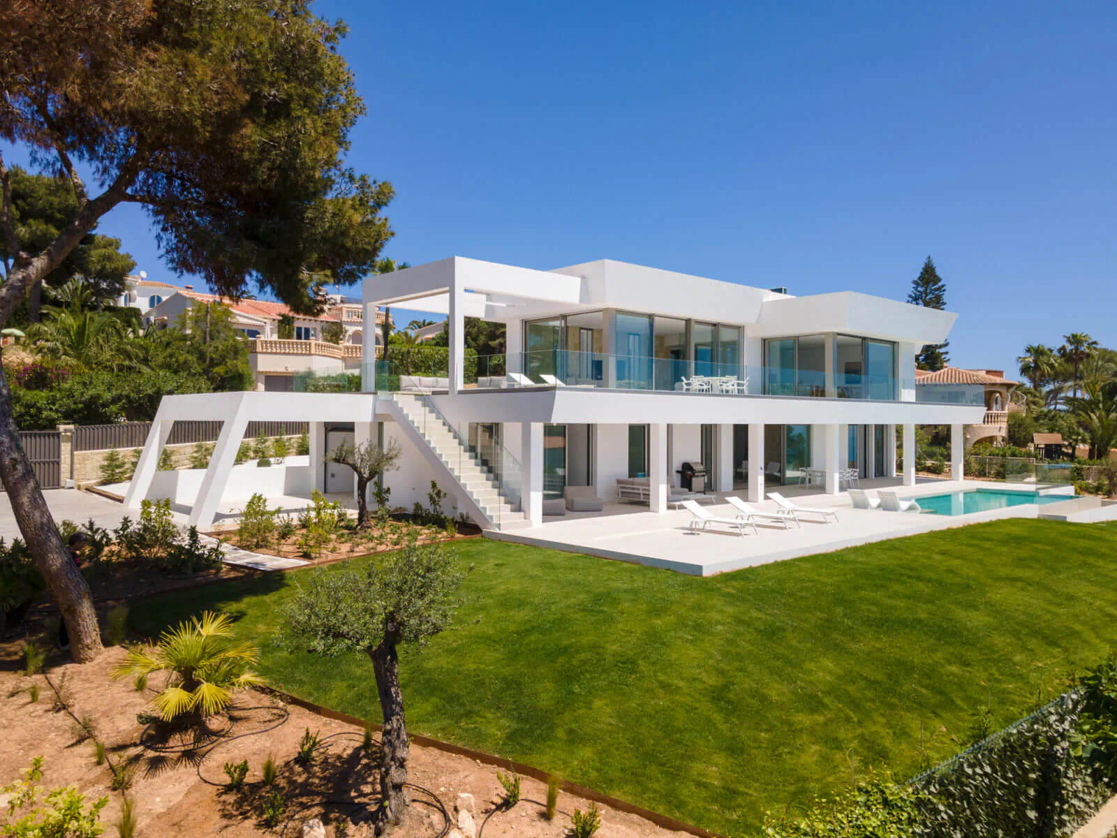 Modern white luxury house with glass walls, infinity pool, lounge chairs, lush green lawn, pine trees, and blue sky backdrop