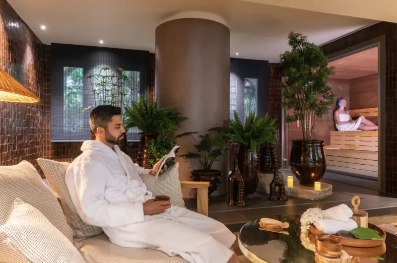 Man in white robe reading on sofa in luxurious spa lounge with plants, sauna, and woman inside, Les Étangs de Corot Paris.