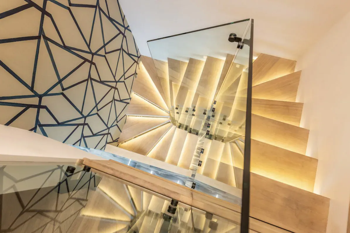 Modern spiral staircase with wooden steps, glass railings, LED lighting, and geometric blue wall backdrop