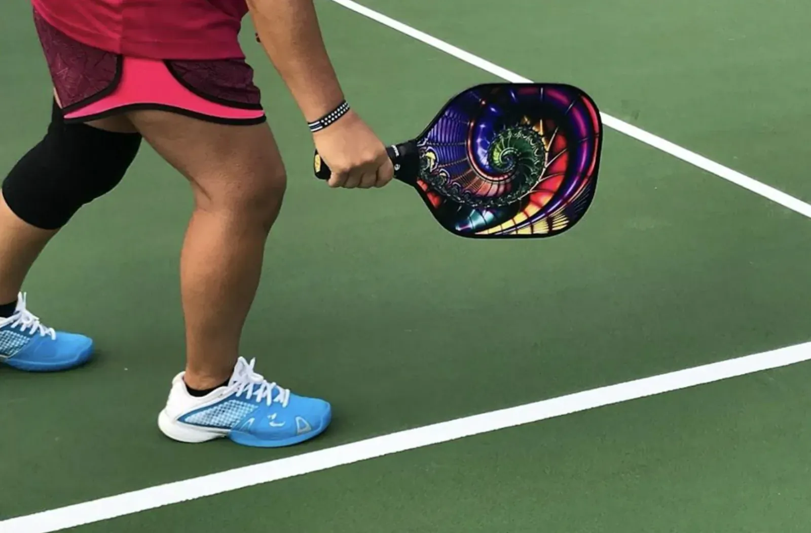 Woman in red shorts and blue shoes holds colorful spiral pickleball paddle on green court, ready to play.
