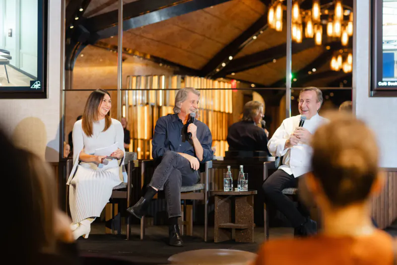 Panel discussion with four professionals on stage: two men speaking into mics, two women seated, modern venue with wood beams and pendant lights.