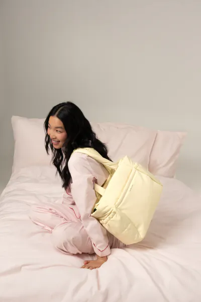 Smiling Asian woman in pink sweatsuit sits on pink bed, wearing yellow Kapten & Son Cloud Club puffy tote bag on back