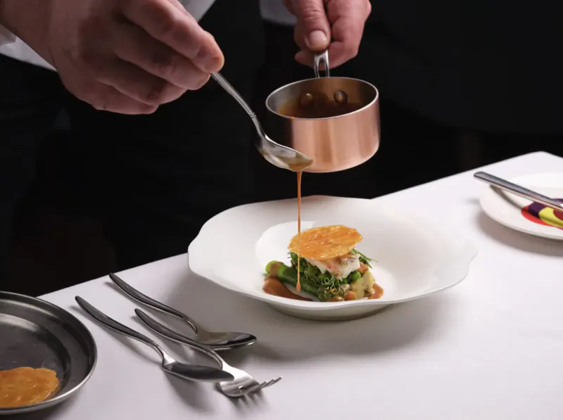 Chef pouring sauce from copper pan onto plated dish with greens and orange crisp in cooking masterclass
