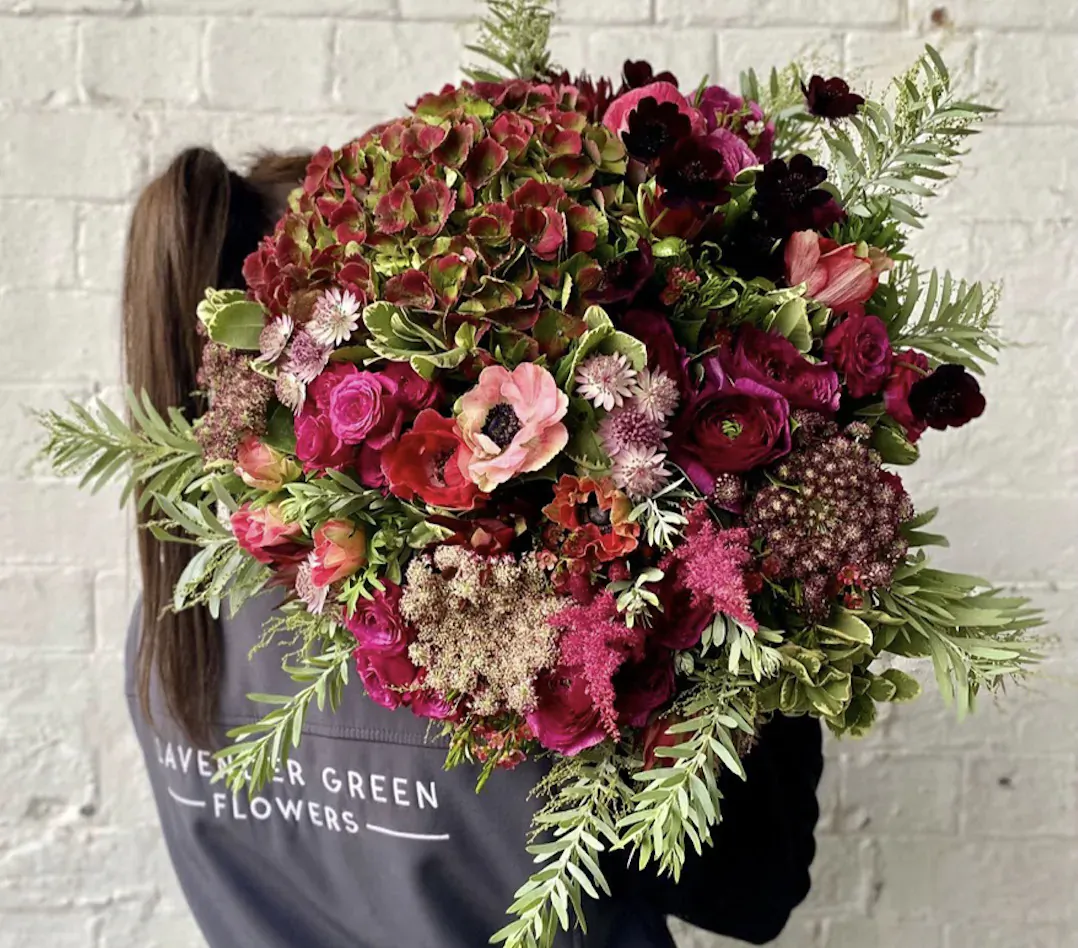 Person from behind holding large bouquet of red hydrangeas, roses, greenery against white brick wall, wearing 'Lavender Green Flowers' shirt