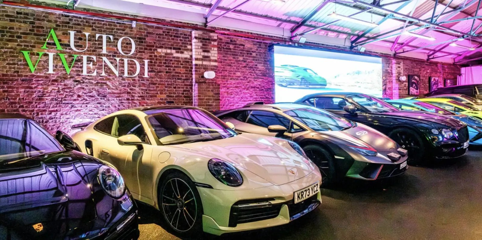 Auto Vivendi showroom with white Porsche 911 GT3 RS, Lamborghinis, and other supercars under pink neon lights against brick wall.