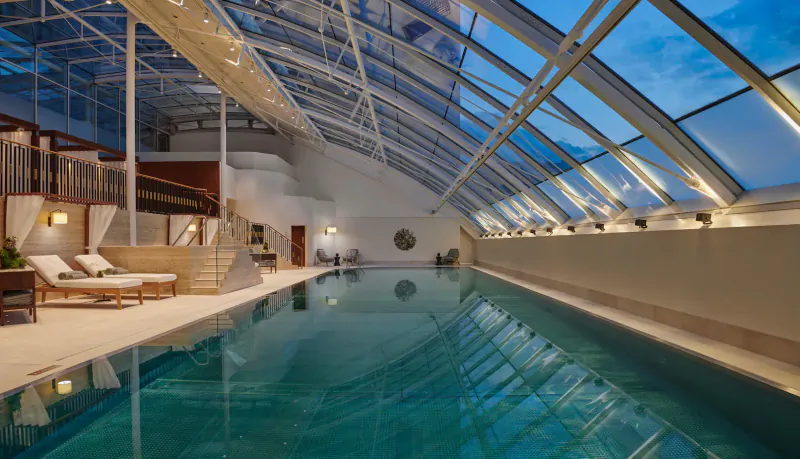 Luxurious indoor pool at dusk under expansive glass atrium with lounge chairs and plants, Carlton Tower Jumeirah.