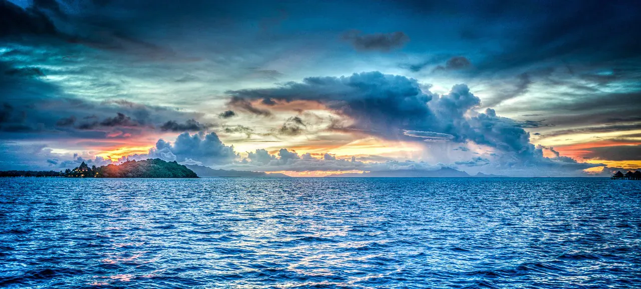 Dramatic sunset over ocean with distant island, dark blue clouds and golden sky glow.