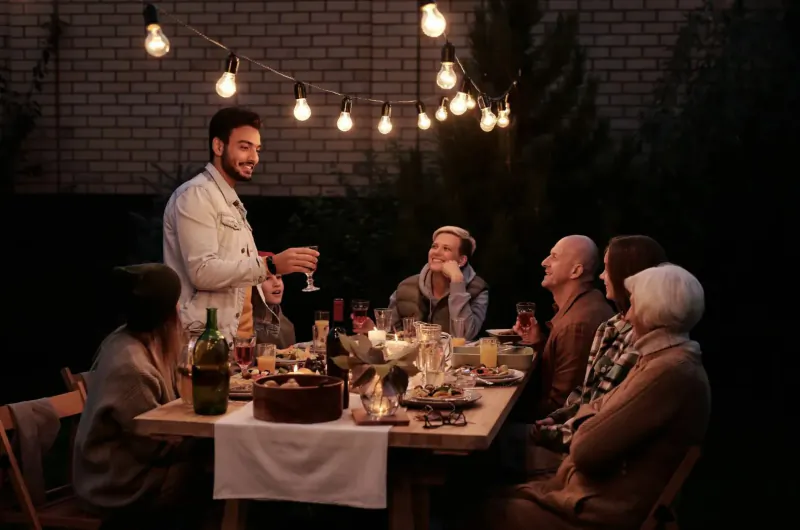 Group of smiling people at outdoor evening dinner table with wine bottles, glasses, cake, and string lights