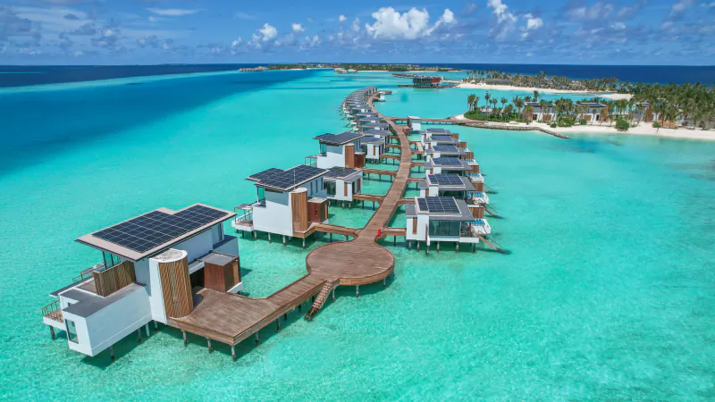 Aerial view of modern overwater villas with solar panels on wooden walkways at turquoise Crossroads Maldives lagoon.