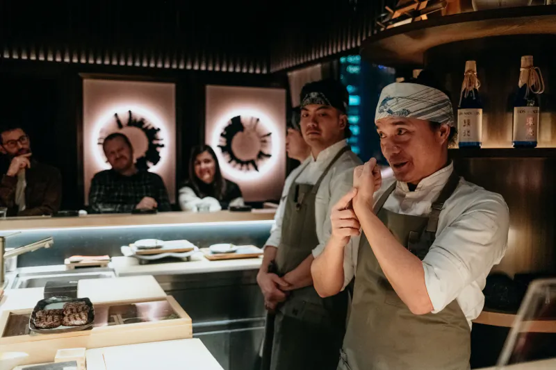 Chef in headband and apron gesturing at counter in LUNA Omakase, with diners, sake bottles, and circular wall art.