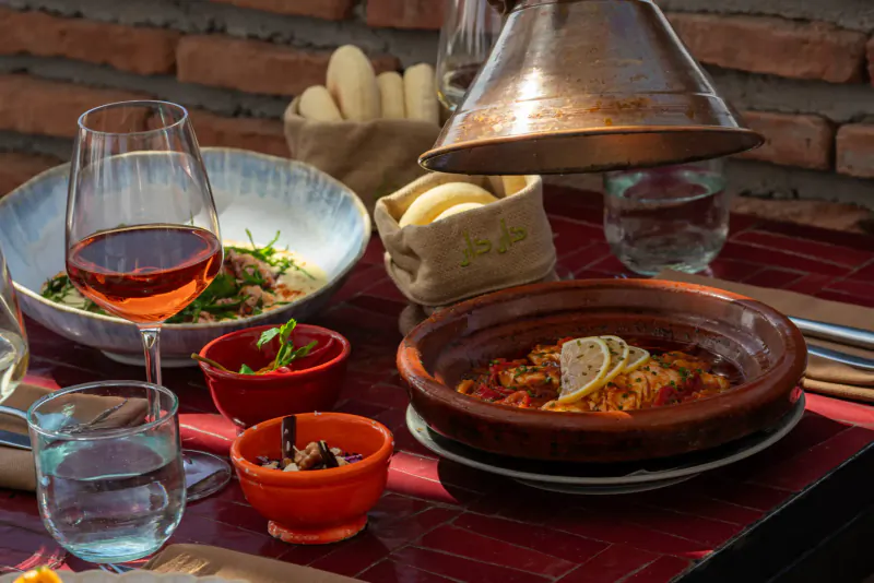 Moroccan rooftop table: tagine with lemon, couscous salad, bread, orange dips, wine, water on red tiles against brick wall