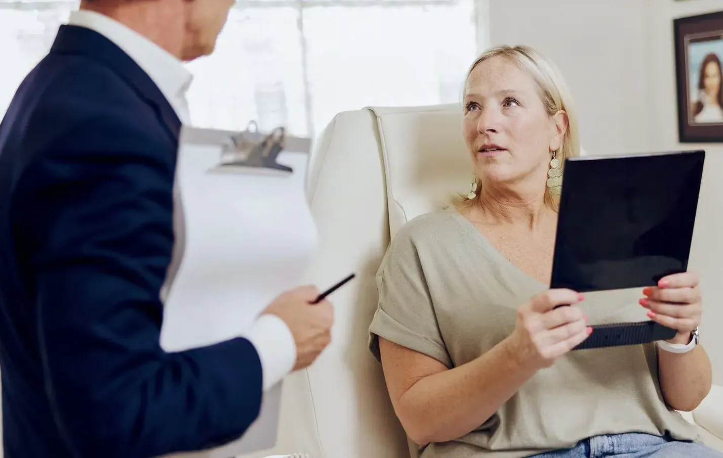 Suit-clad man discusses with surprised blonde woman holding tablet in clinic, pointing at papers