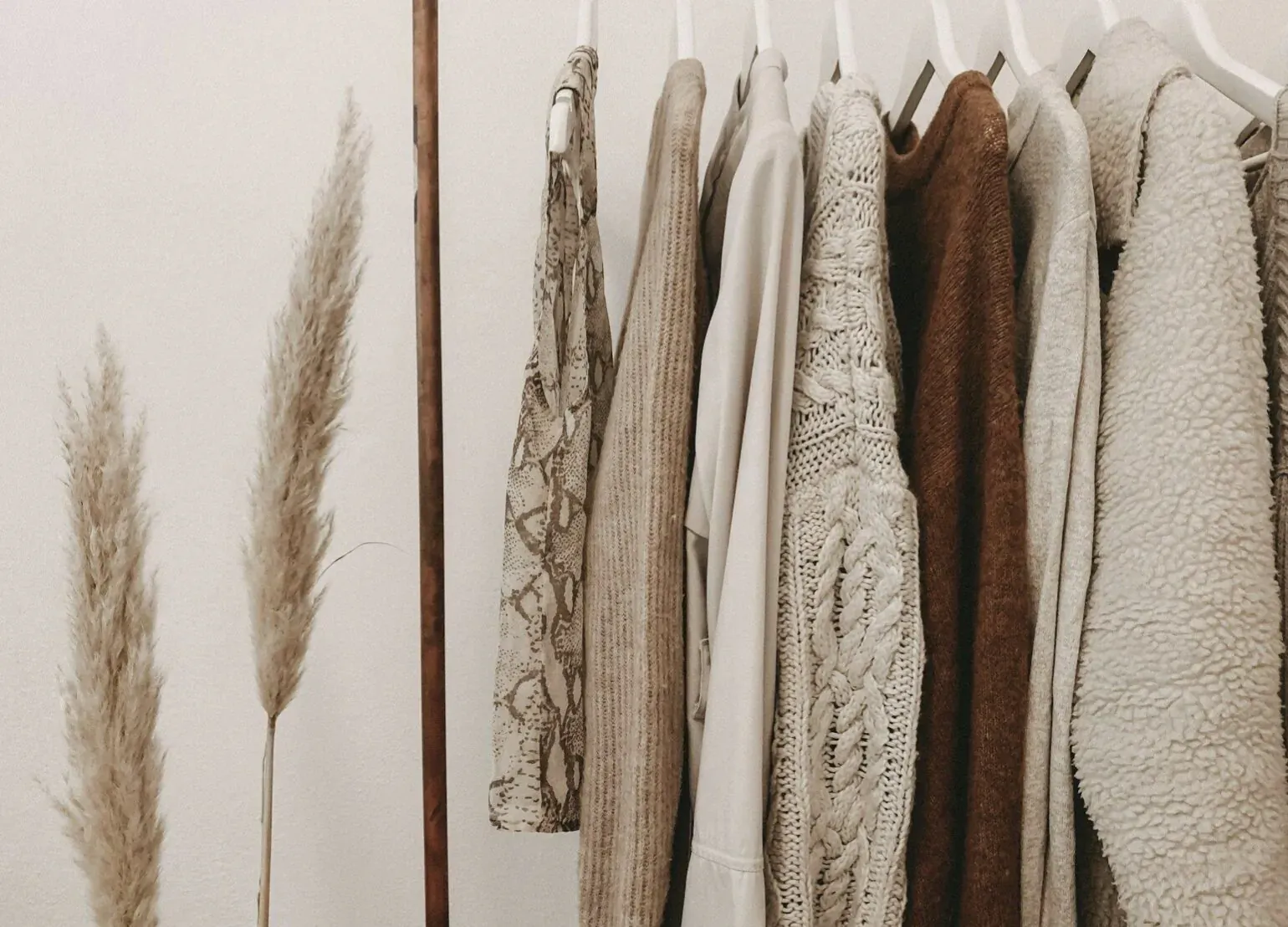 Assorted chic sweaters and knits hanging on wooden rack with pampas grass decoration
