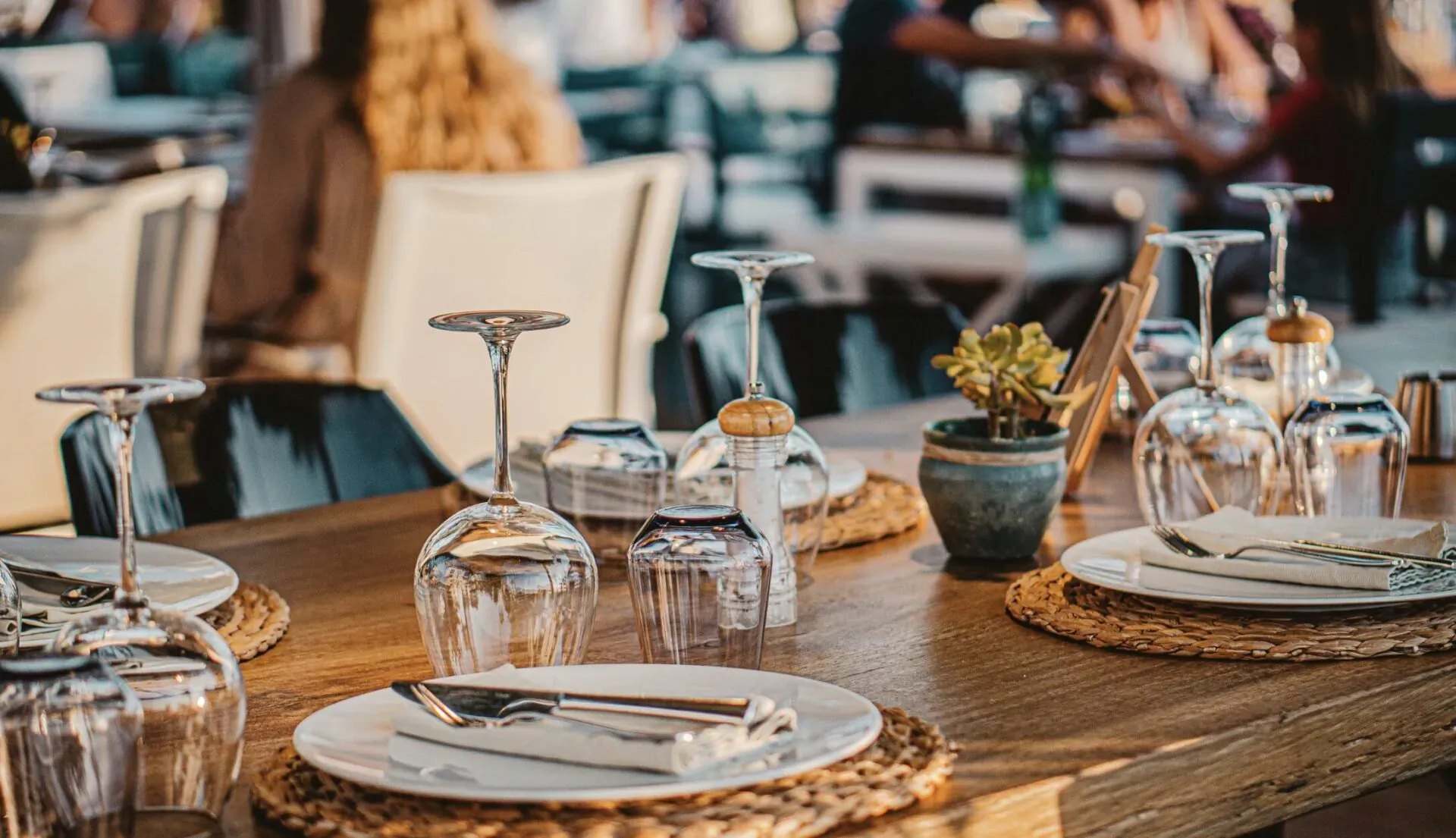 Elegant fine dining table set with wine glasses, plates, napkins, and small plant on woven placemats, outdoor blurred background
