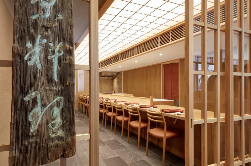 Entrance to Roketsu kaiseki restaurant with wooden sign 'Roketsu' in turquoise kanji, shoji screens, long wooden counter, chairs, and warm lighting.