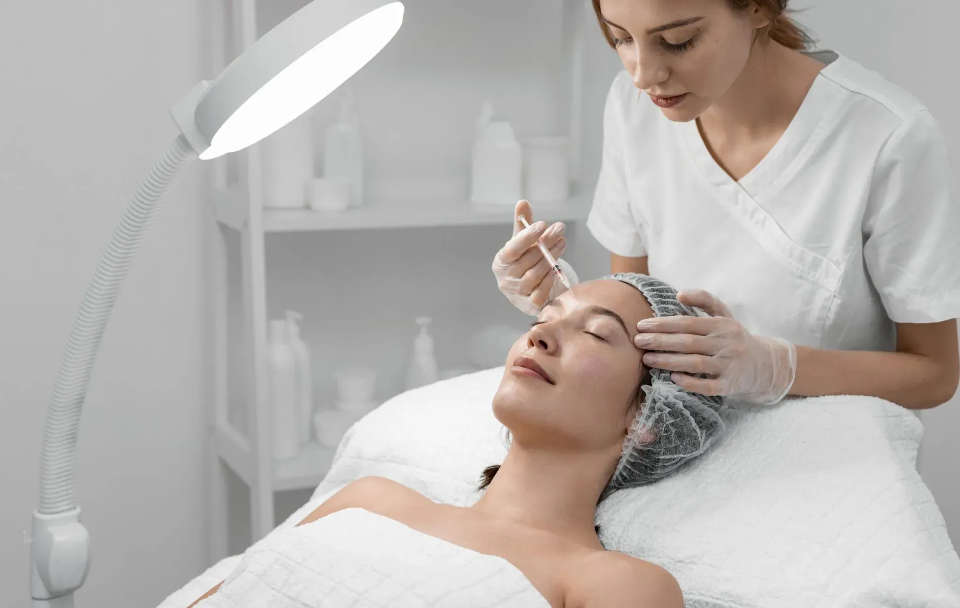 Nurse in white uniform injecting Botox into reclined woman's forehead in clinic under bright lamp, both wearing hairnets and gloves.