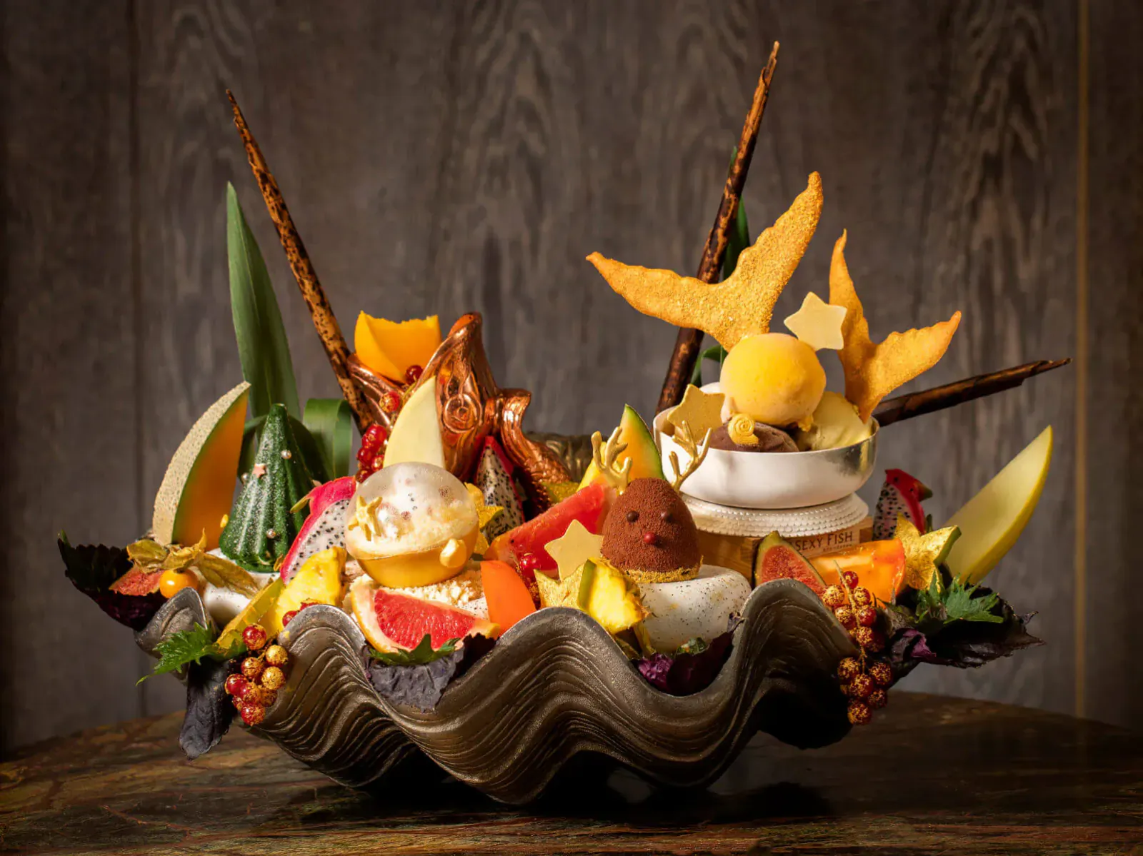 Vibrant fruit platter in giant seashell, garnished with star-shaped pastries, custards, and festive elements on wooden surface.