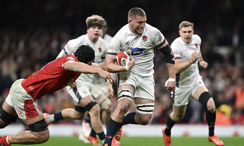 England rugby player in white sprints with ball, tackled by red Wales player, teammates support on stadium pitch