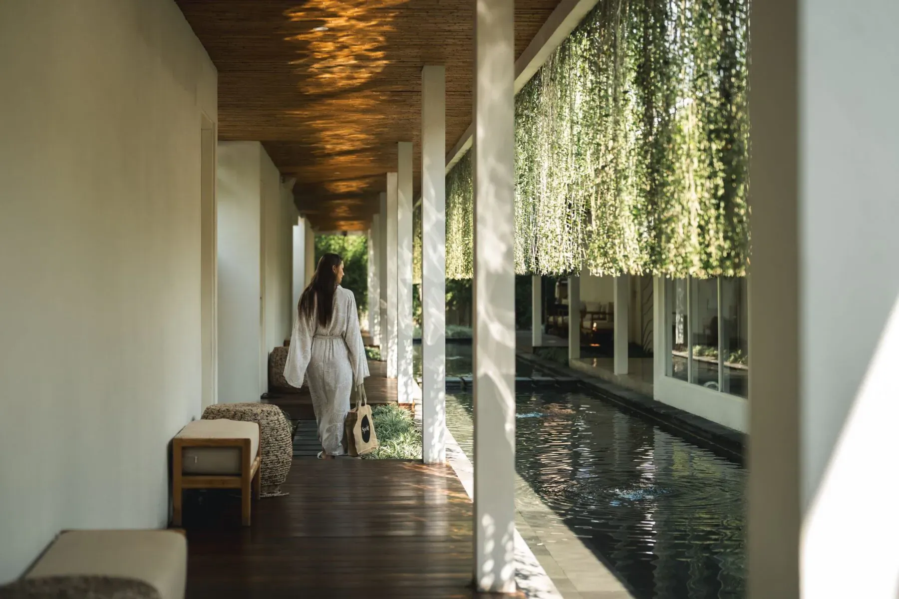 Woman in white walking down luxurious Bali spa hallway with wooden pillars, hanging vines, and reflecting pool