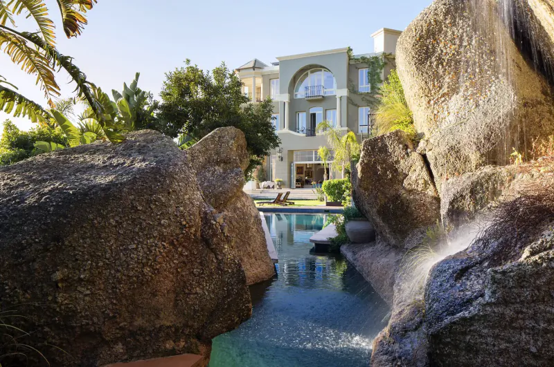 Luxury Cape Town home framed by palm trees and boulders, with infinity pool overlooking ocean view.
